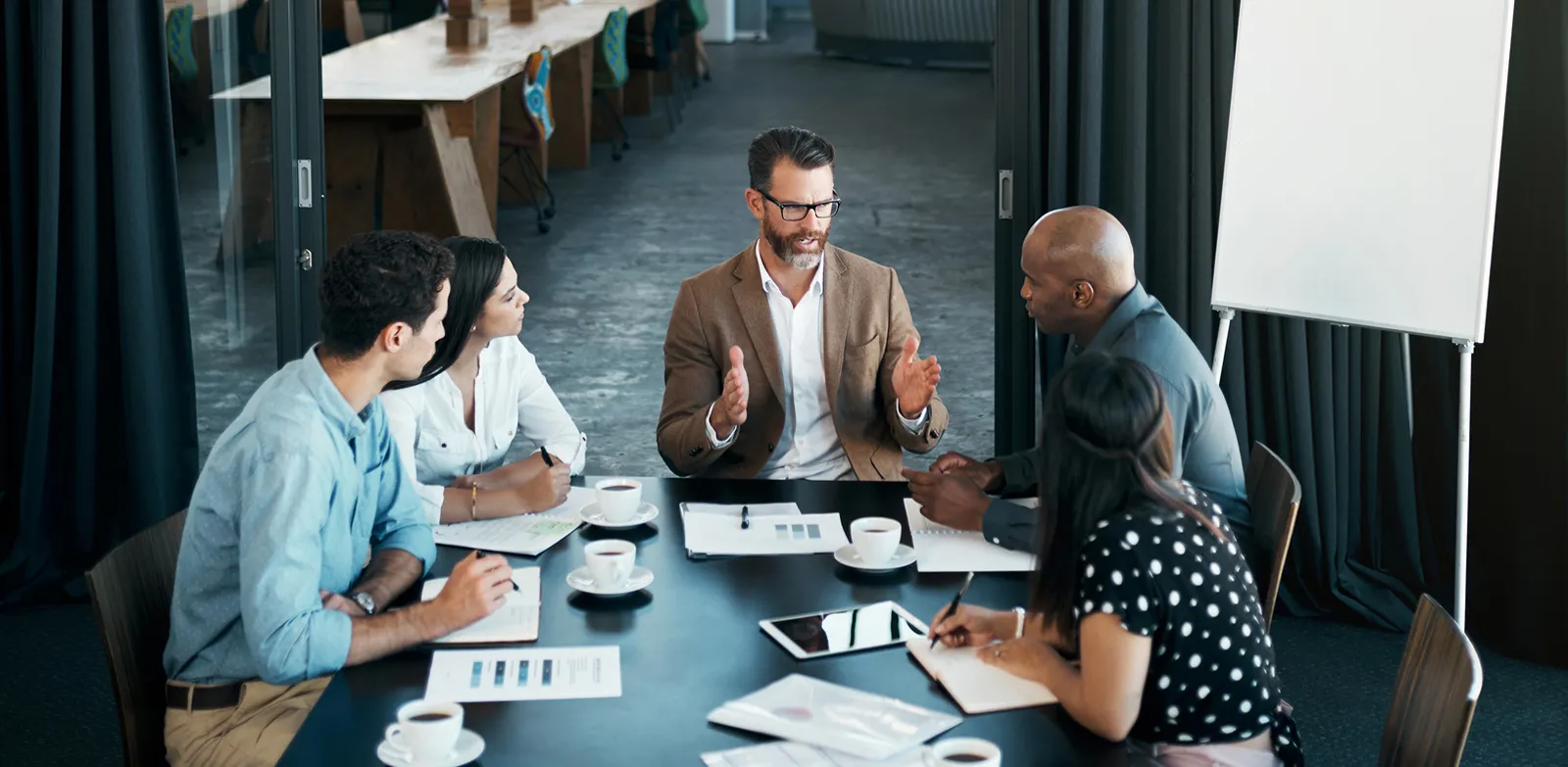 Five people sit around a large table scattered with cups of coffee and working documents.